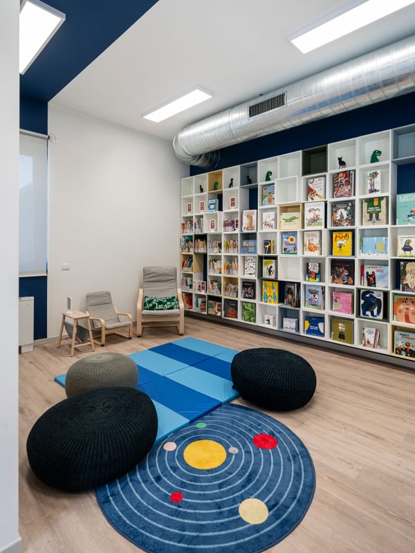 Un’aula studio della Biblioteca comunale di Brescia Roncadelle con una combinazione di mobili da parete EKET in cui all'interno sono esposti volumi dalle copertine colorate. Al centro della stanza ci sono un tappeto AFTONSPARV e, insieme ai pouf SANDARED e ai tappetini PLUFSIG e due poltrone POÄNG. La stanza è illuminata da pannelli a LED da soffitto.