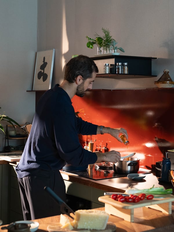 Un uomo ai fornelli versa un filo d'olio in una pentola, circondato da ingredienti freschi come pomodorini e formaggio.