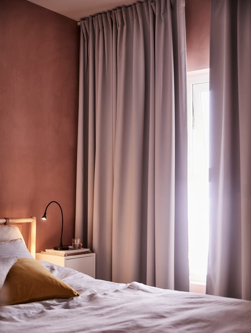 Two full-length MAJGULL curtains hang in a window, partly open to let light into a bedroom with a bed and bedside table.