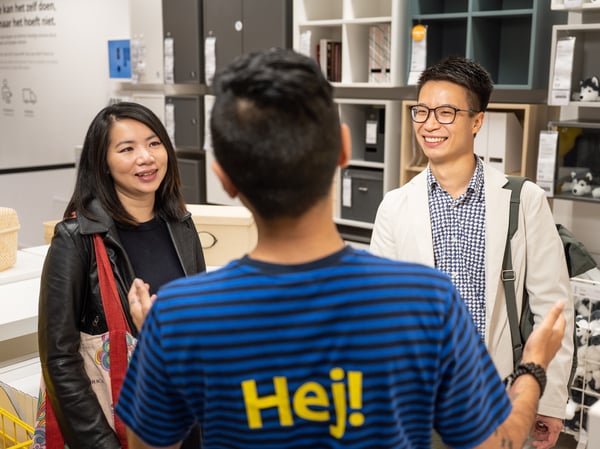 IKEA employee greeting two customers inside a store showroom, with shelving units displayed in the background.