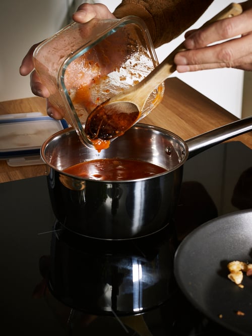 Close up on induction hob with a person pouring the contents of a glass food container into a stainless steel saucepan.