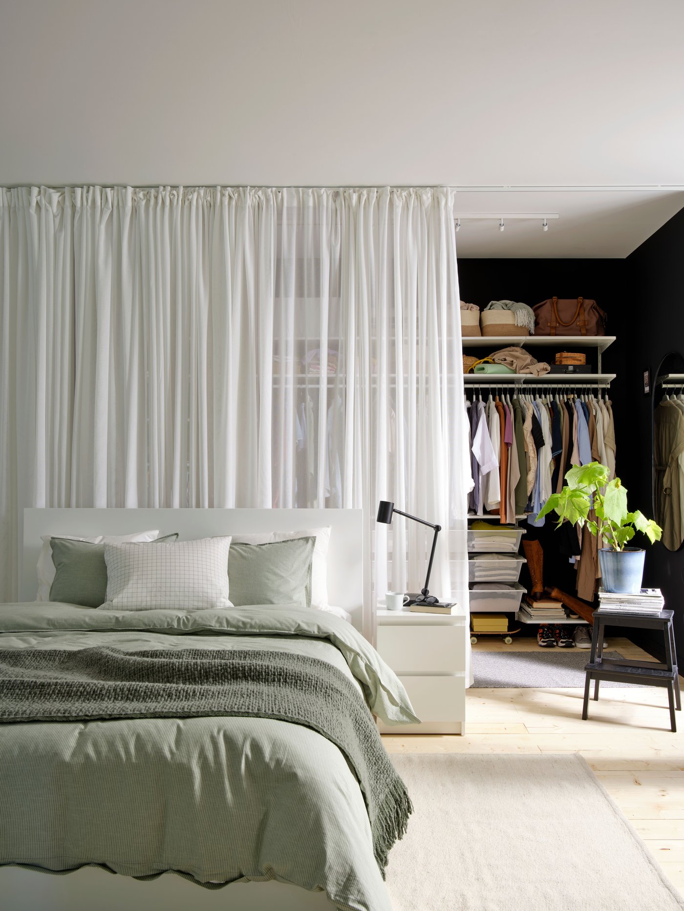 Sovrum med ljusgrönt bäddset och sängbord, och bakom ett vitt draperi syns en öppen garderob med kläder, hyllor och en växt på en pall.