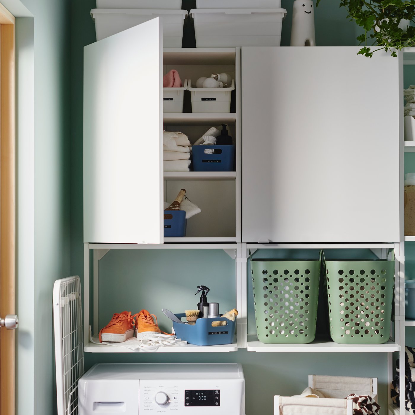 An open, grey-blue UPPDATERA box holds shoe cleaning tools by a pair of shoes on a deep wall shelf above a washing machine.