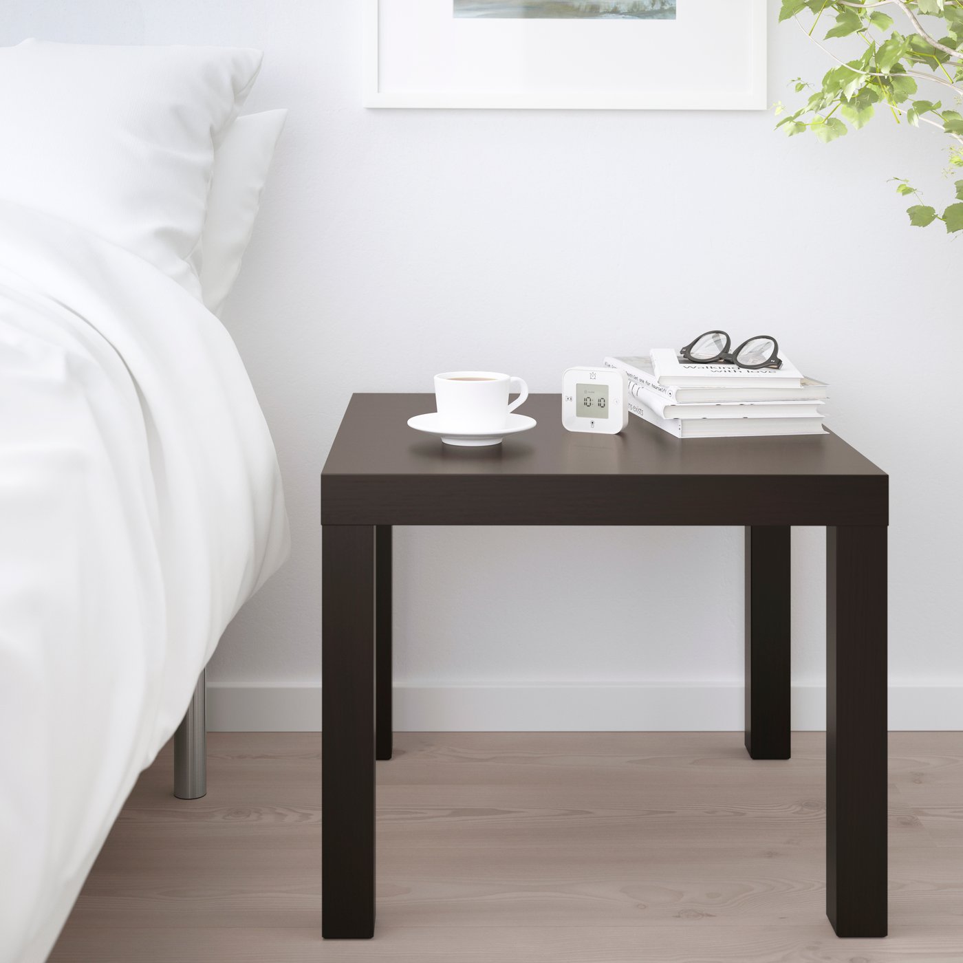 Ein schwarzbraunner LACK Beistelltisch neben einem weiß bezogenen Bett ist mit einer Tasse, einem kleinen Wecker, mehreren Büchern und einer Brille dekoriert.