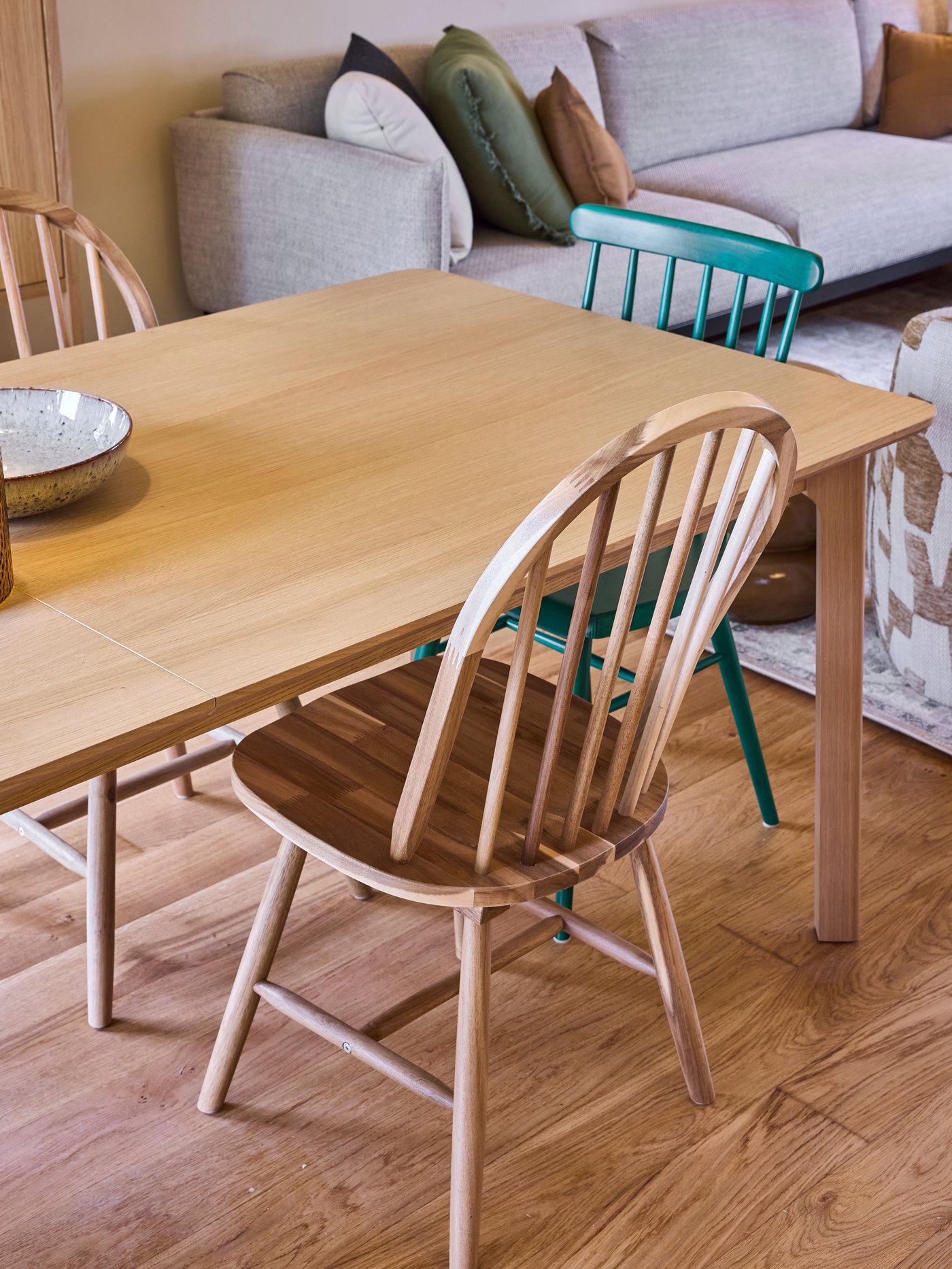 Houten eettafel met mix van stoelen op houten vloer, met sofa en kussens op de achtergrond in een warme, huiselijke leefruimte.