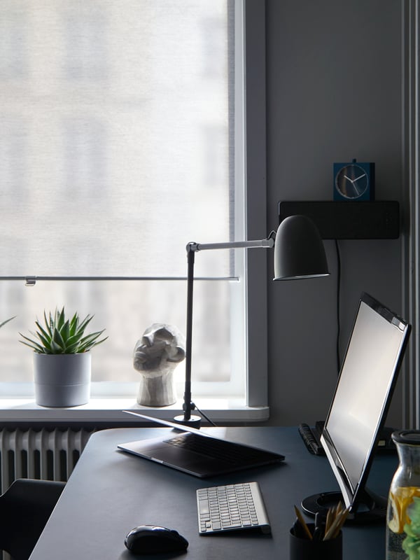 Window with SKOGSKLÖVER grey roller blind drawn almost down, behind a desk with a lamp and computer screen.