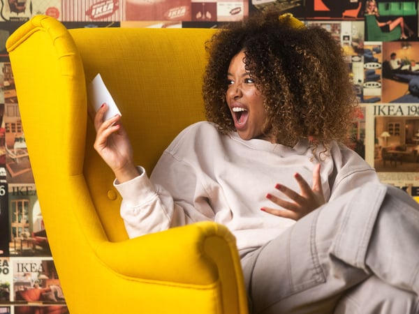 A woman with curly hair, wearing a light-colored sweatshirt and pants, sits in a yellow armchair and looks surprised at her phone.