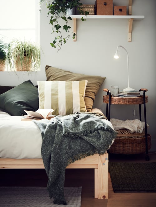 Cameră de zi cu un cadru de pat cu mai multe perne și o aruncătură, plasat lângă o masă cu cărucior cu depozitare și o lampă de lucru.