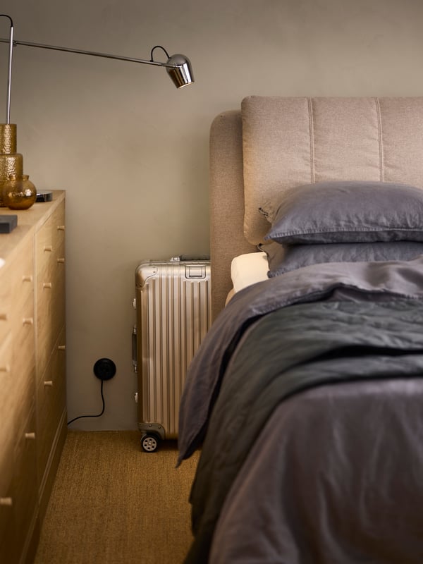 Due cassettiere TONSTAD impiallacciate in rovere con 6 cassetti di fronte a un letto imbottito LILLFJÄLLET con una valigia dietro la testata.