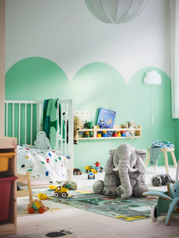 A toddler’s room with green/white walls, GULLIVER cot, VALLABY rug and JÄTTESTOR soft toy elephant on the floor.