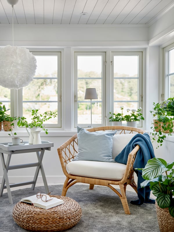 Ljus hörna i ett rum med stora fönster, STOCKHOLM rottingfåtölj med vita dynor och blå kuddar, grått sidobord, gröna krukväxter i fönstret, öppen bok på rund flätad sittpuff och vit fluffig taklampa.