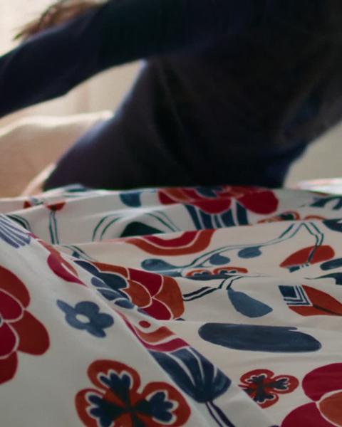 Mulher cai pesadamente sobre cama de casal com estrutura em aço em tons escuros, enquanto a roupa de cama muda entre cores diferentes e padrões florais.