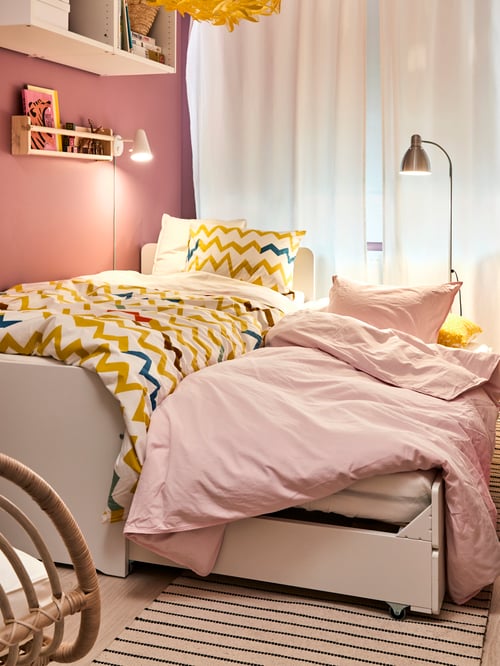 Une chambre d’enfant avec une structure de lit blanche et un dessous de lit allongé, deux ensembles de literie, un rangement mural, une applique et un tapis rayé.