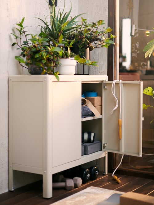 Une armoire KOLBJÖRN beige avec une porte ouverte et des boîtes sur des étagères à l’intérieur. Des plantes sont posées dessus et des poids se trouvent en dessous.