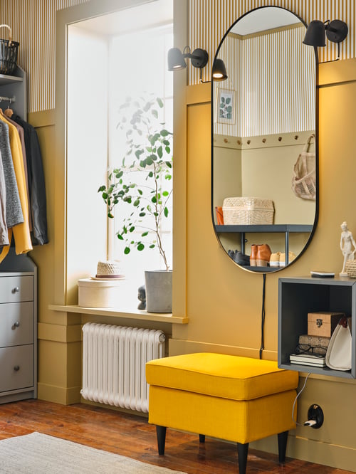 Bright hallway with large oval mirror with gold rim on the wall, yellow footstool, wall shelf, open wardrobe with drawers.