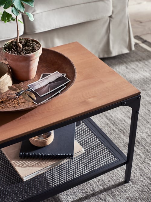 A black and pine FJÄLLBO coffee table is on a rug by a sofa. It holds notebooks on its shelf with a tray of photos on top.