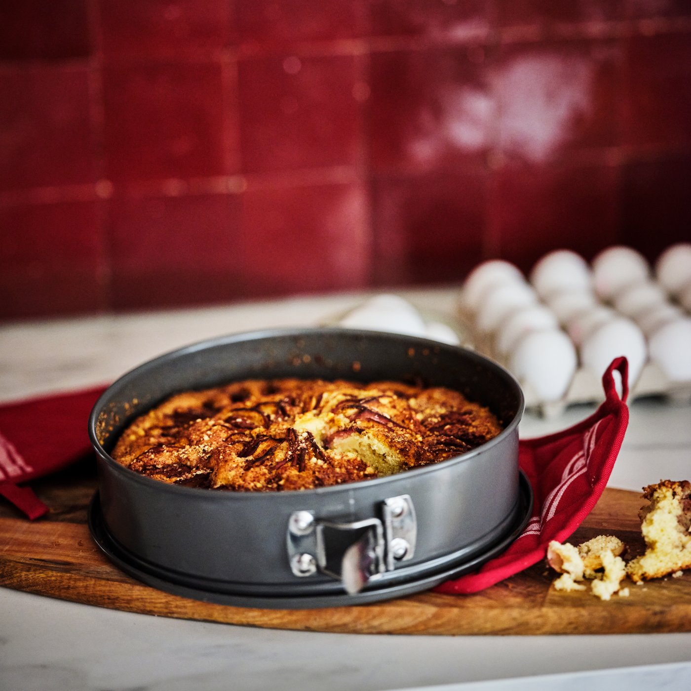 A worktop in a red-tiled kitchen where a freshly baked pie in a grey HEMMABAK springform rests on red HILDEGUN pot holders.