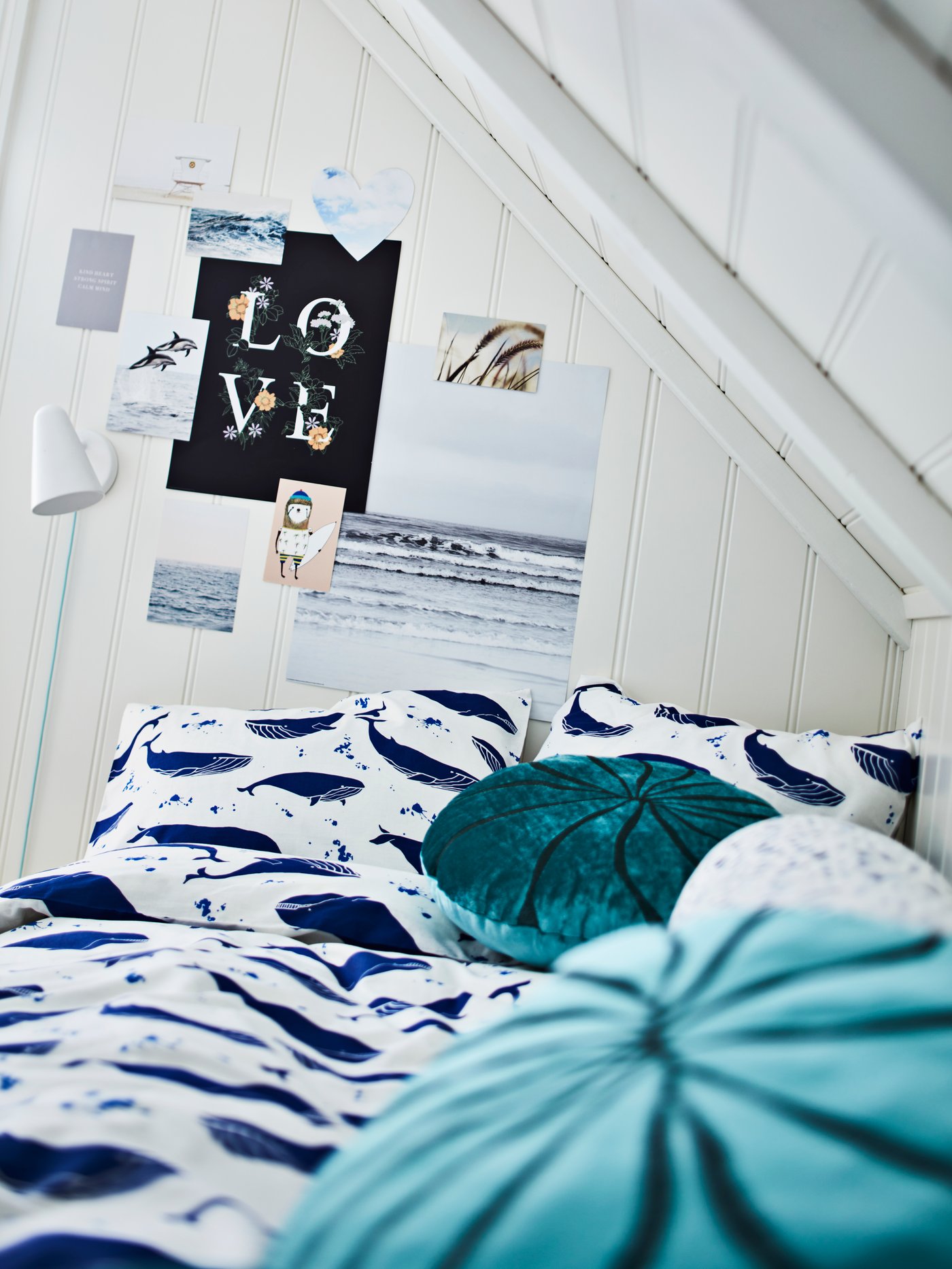 A bed with BLÅVINGAD bed linen and two turquoise BLÅVINGAD cushions stands in a corner in a room with a sloping ceiling.
