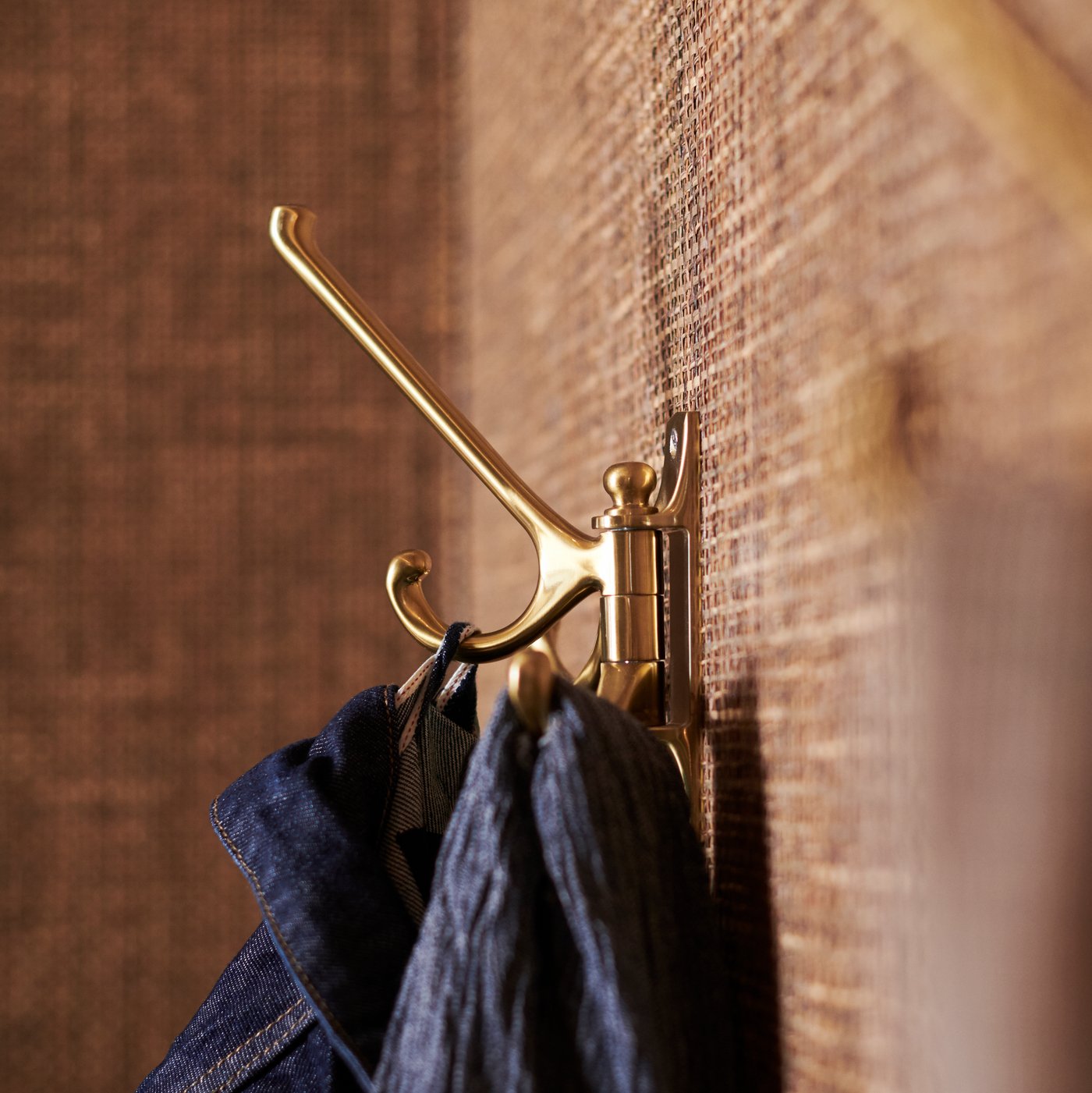 A brass-colour KÄMPIG three-arm hook attached to a light brown sea-grass wall. It holds a denim jacket and a dark blue scarf.