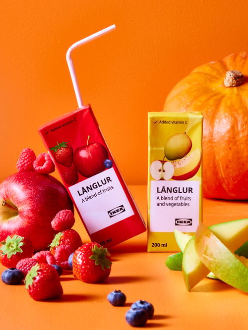 Two LÅNGLUR fruit smoothies, one red and one yellow sit on an orange surface along with multiple fruit items.