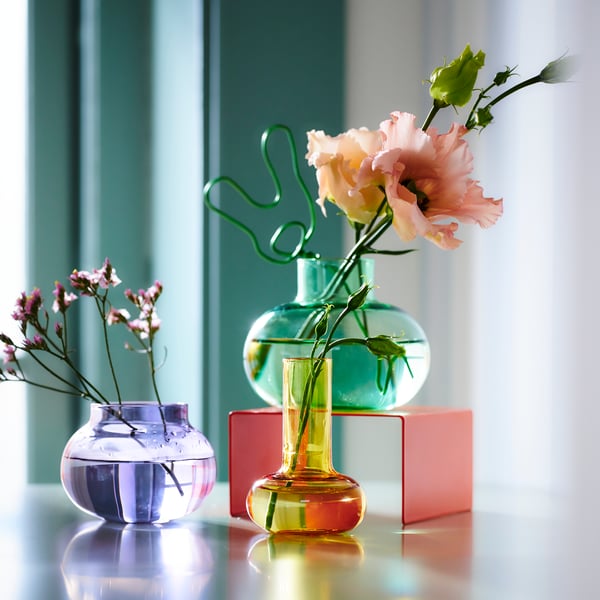 Drei bunte TJÄRLEK Vasen stehen mit einzelnen Blumen dekoriert auf einem Tisch.