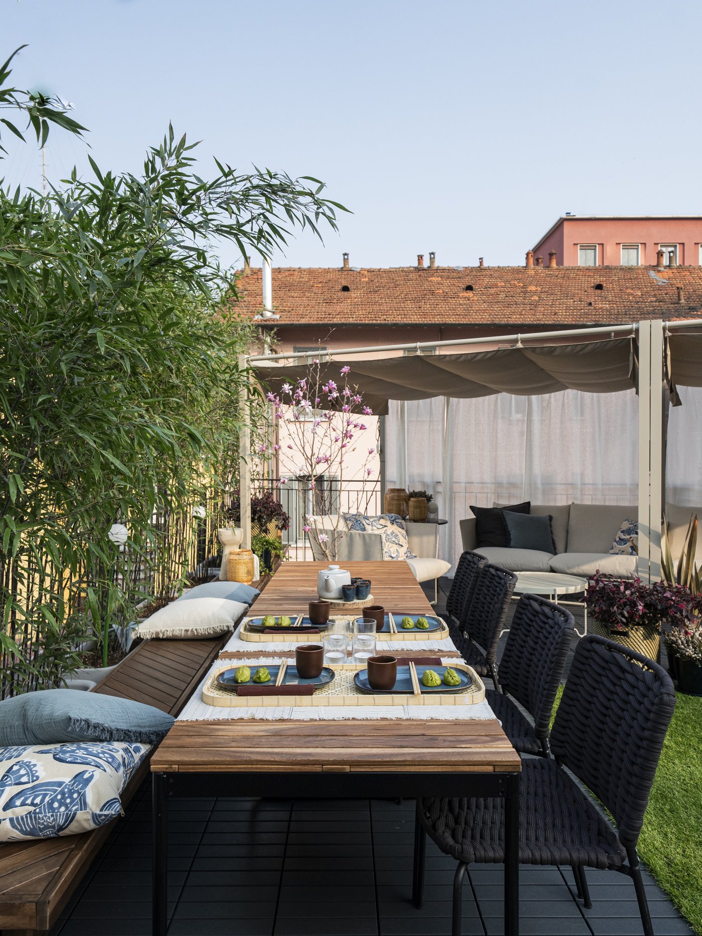 Terrazza con un tavolo VÄSMAN allungabile in legno marrone chiaro, apparecchiato e con sedie nere intrecciate accanto, circondato da piante verdi.