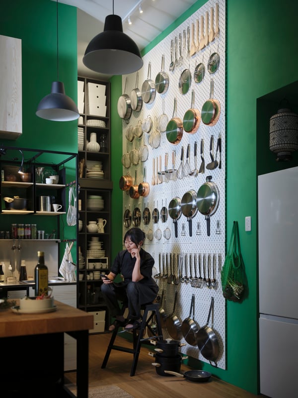 A person is sitting on a stepladder in front of a wall adorned with SKÅDIS pegboards, with kitchen items attached to them.