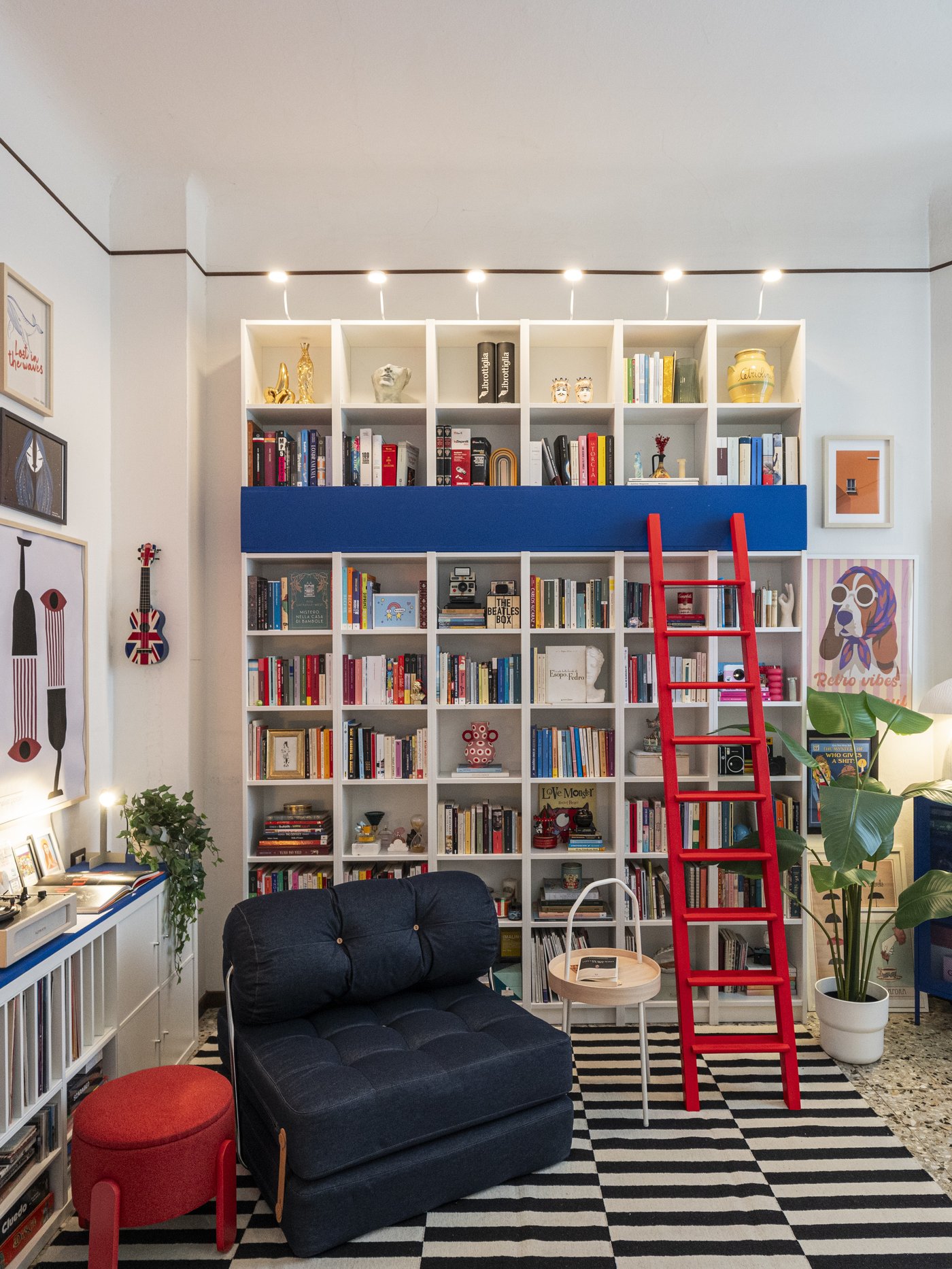 Soggiorno colorato con grande libreria BILLY bianca a parete piena di libri e oggetti decorativi, scala rossa appoggiata e poltrona blu jeans su tappeto a righe in primo piano.