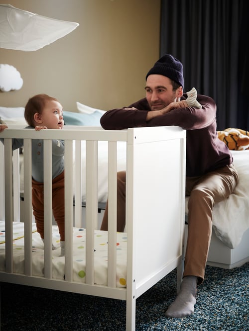Un bărbat ținând în mână o jucărie de pluș se uită la un copil care stă în picioare într-un pătuț SUNDVIK alb.
