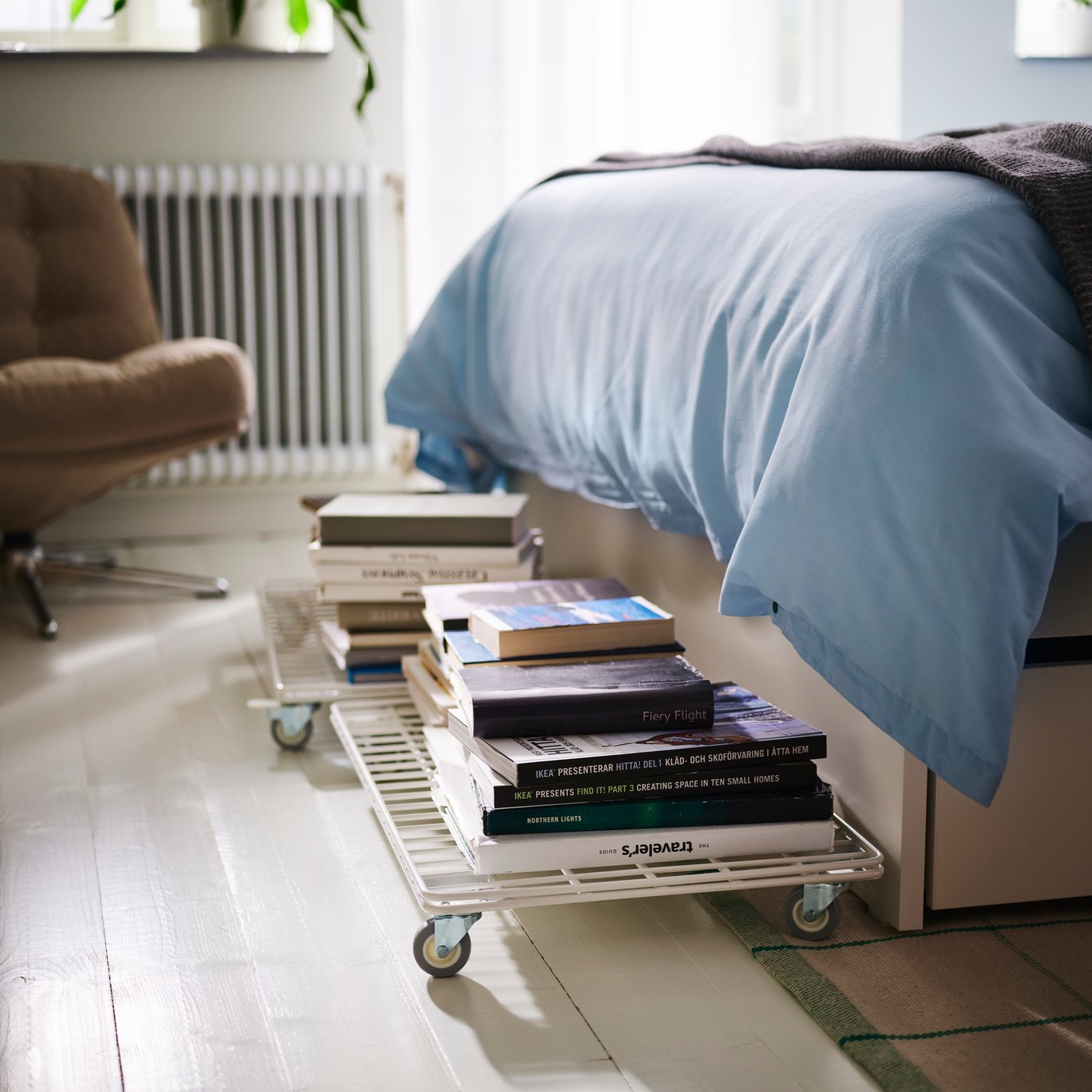 An INVALLNING trolley holding stacks of books on the bedroom floor beside a bed, armchair, window and radiator.