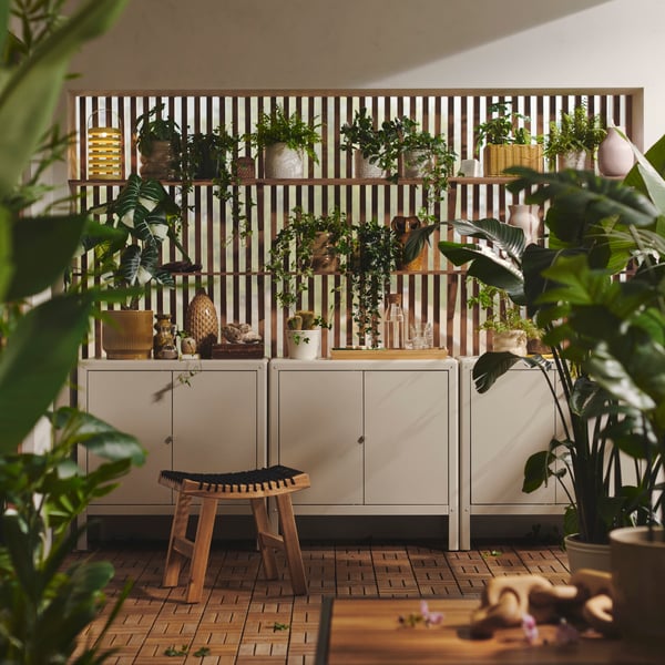 A bright balcony with three beige KOLBJÖRN cabinets, wooden wall shelves displaying various plants on a wall behind it.
