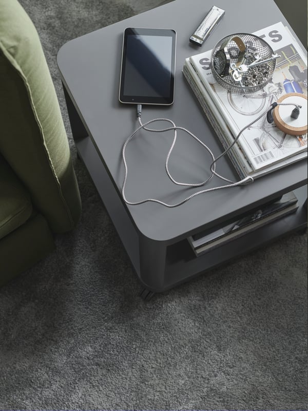 Ein LÖRBY USB-Ladegerät magnetisch auf der Rückseite eines grauen Sofas. An ihm sind zwei Ladekabel befestigt, eines von ihnen führt zu einem Smartphone.