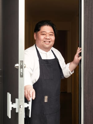 Person in a dark apron standing at an open front door, holding the handle, with warm indoor lighting visible behind.
