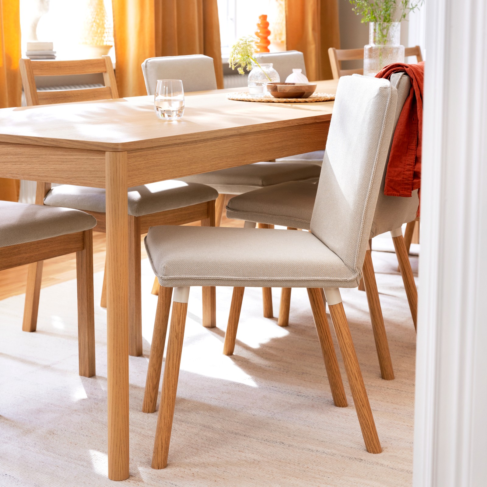 A light-toned TONSTAD chair frame with a white seat is positioned at a wooden dining table, surrounded by other chairs.