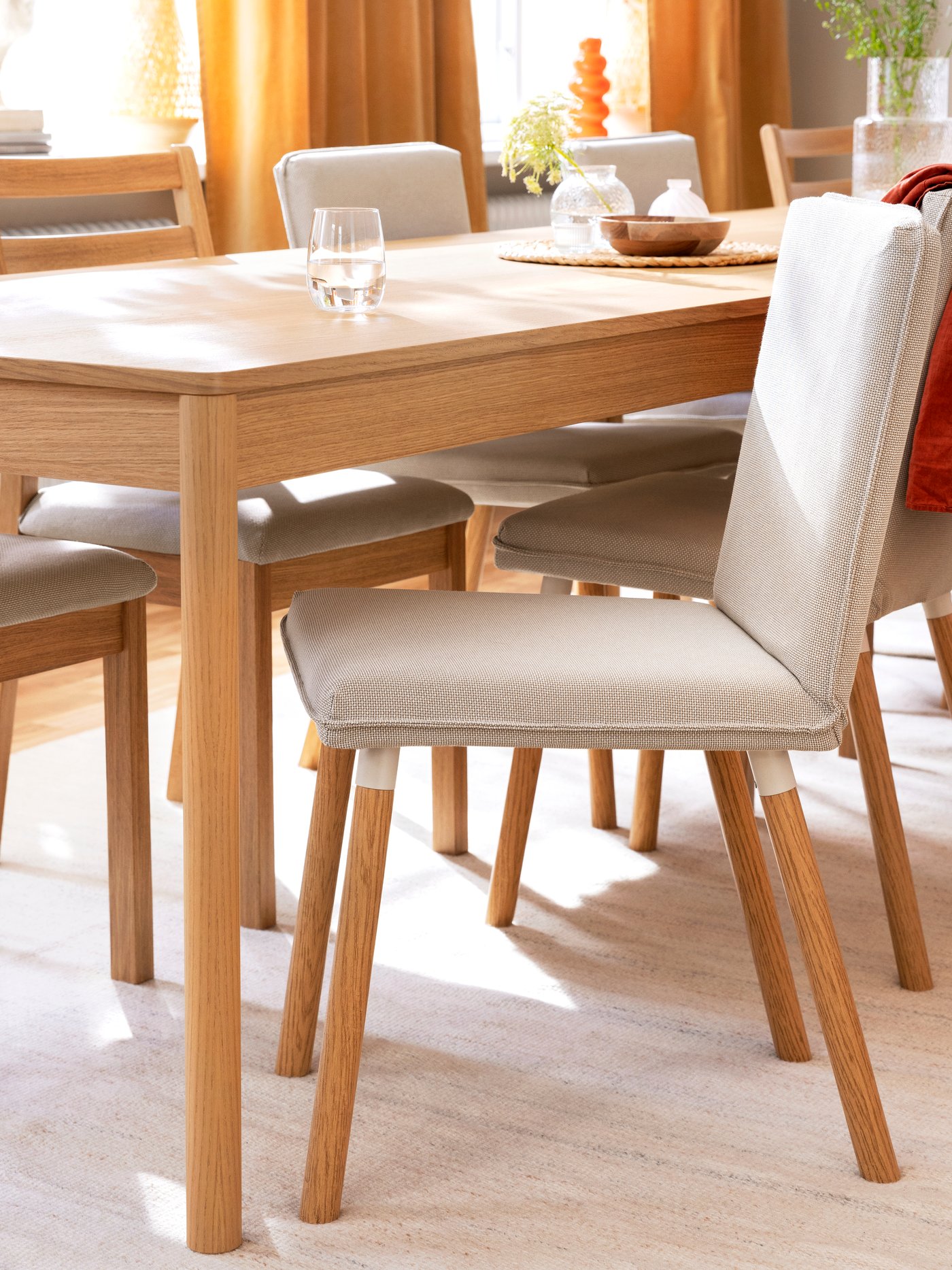 A light-toned TONSTAD chair frame with a white seat is positioned at a wooden dining table, surrounded by other chairs.