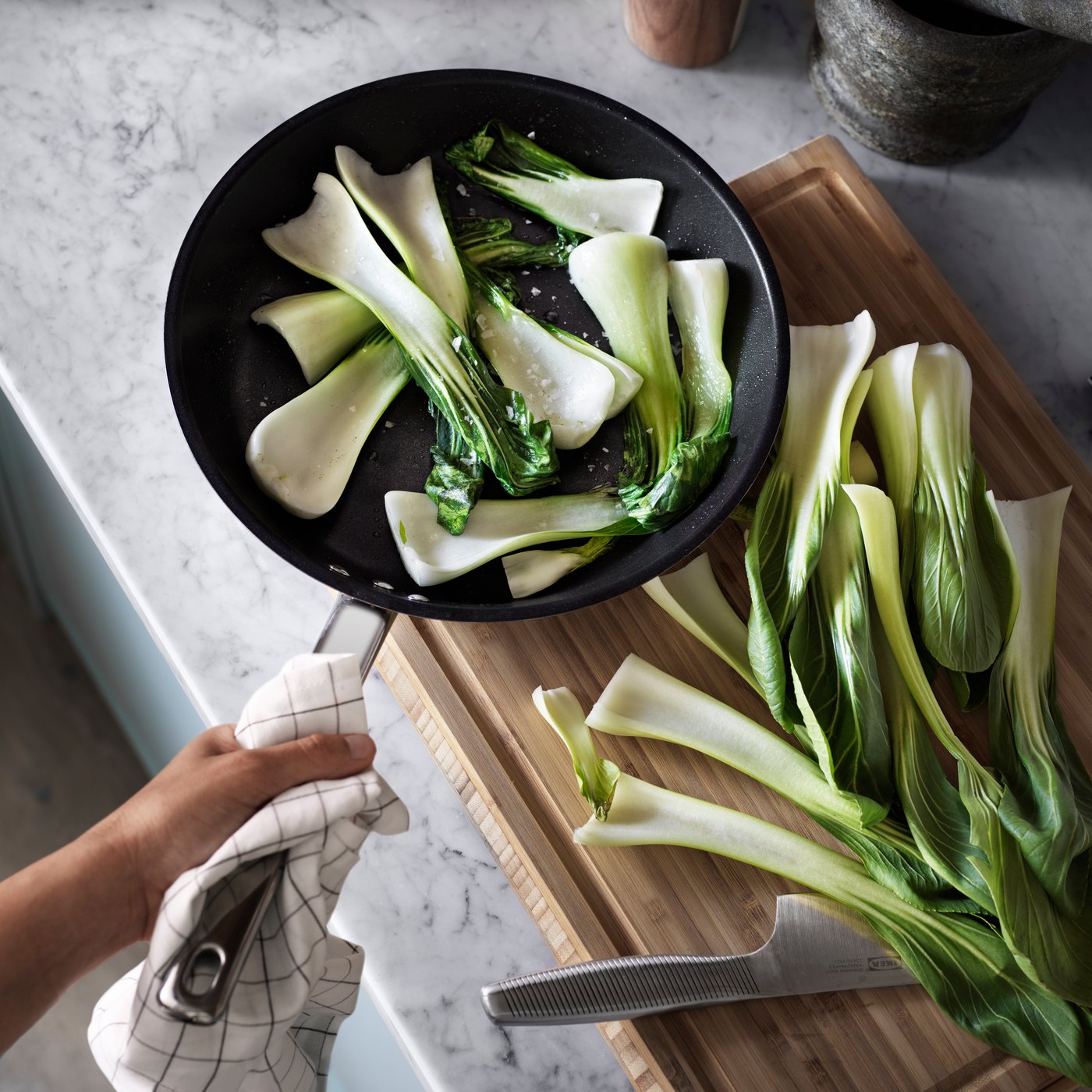 Hand holding a pan with sliced bok choy cooking beside more bok choy on a cutting board with a knife.
