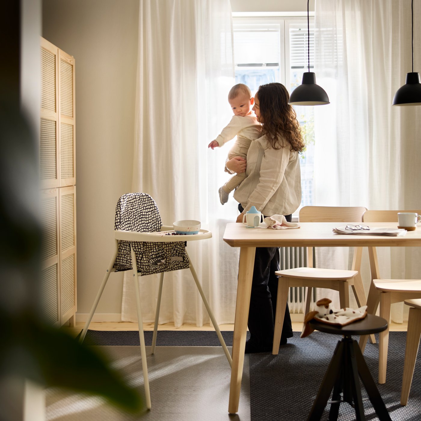 Ein SANDKATT Kinderhochstuhl mit Tablett steht neben einem Esstisch in einem hellen Raum. Daneben steht eine erwachsene Person mit einem Baby auf dem Arm.