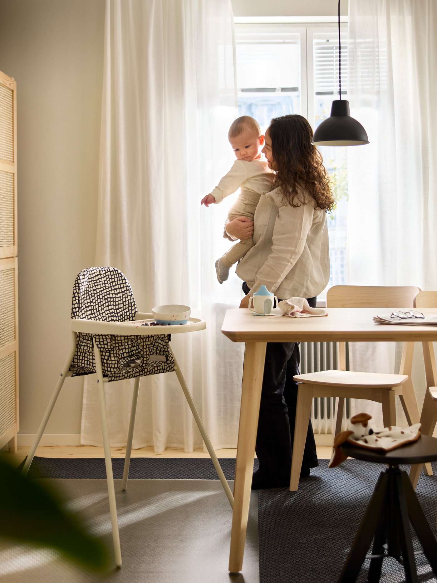 SANDKATT highchair placed beside a dining table in a bright room where an adult is holding a baby.