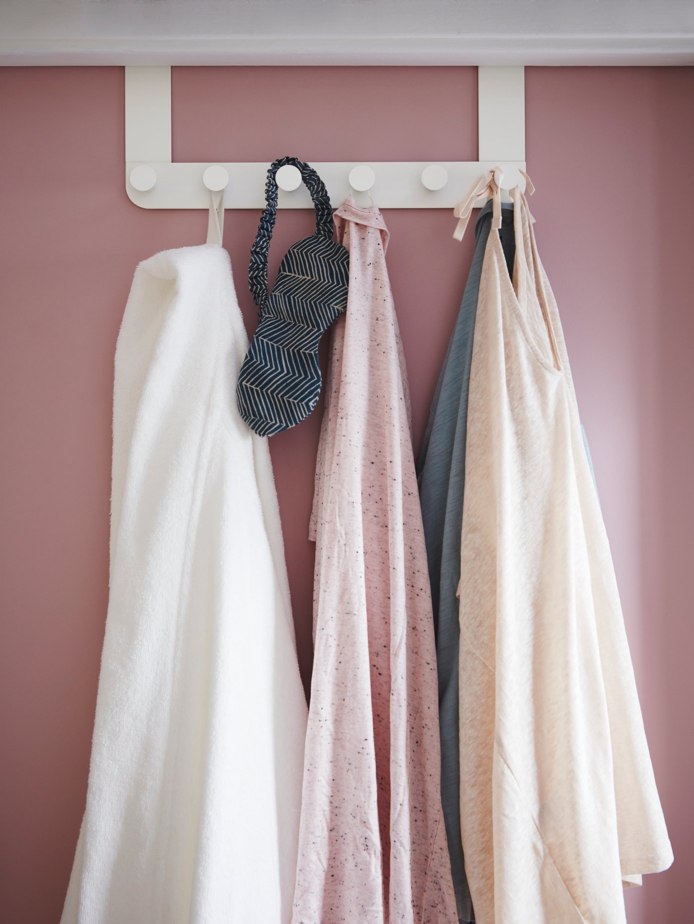 Puerta rosa con perchero de acero blanco con pomos de plástico con una bata blanca, ropa y antifaz para dormir.