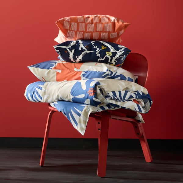 An easy chair sitting by a red wall with a duvet and pillow in HYLSNEJLIKA bedlinen stacked on it, and two cushions on top.