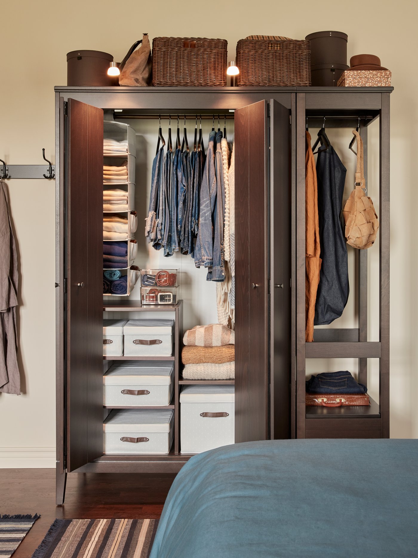 A wooden wardrobe with bi-folding doors open showing white boxes, a hanging shelf organiser and jeans on hangers.
