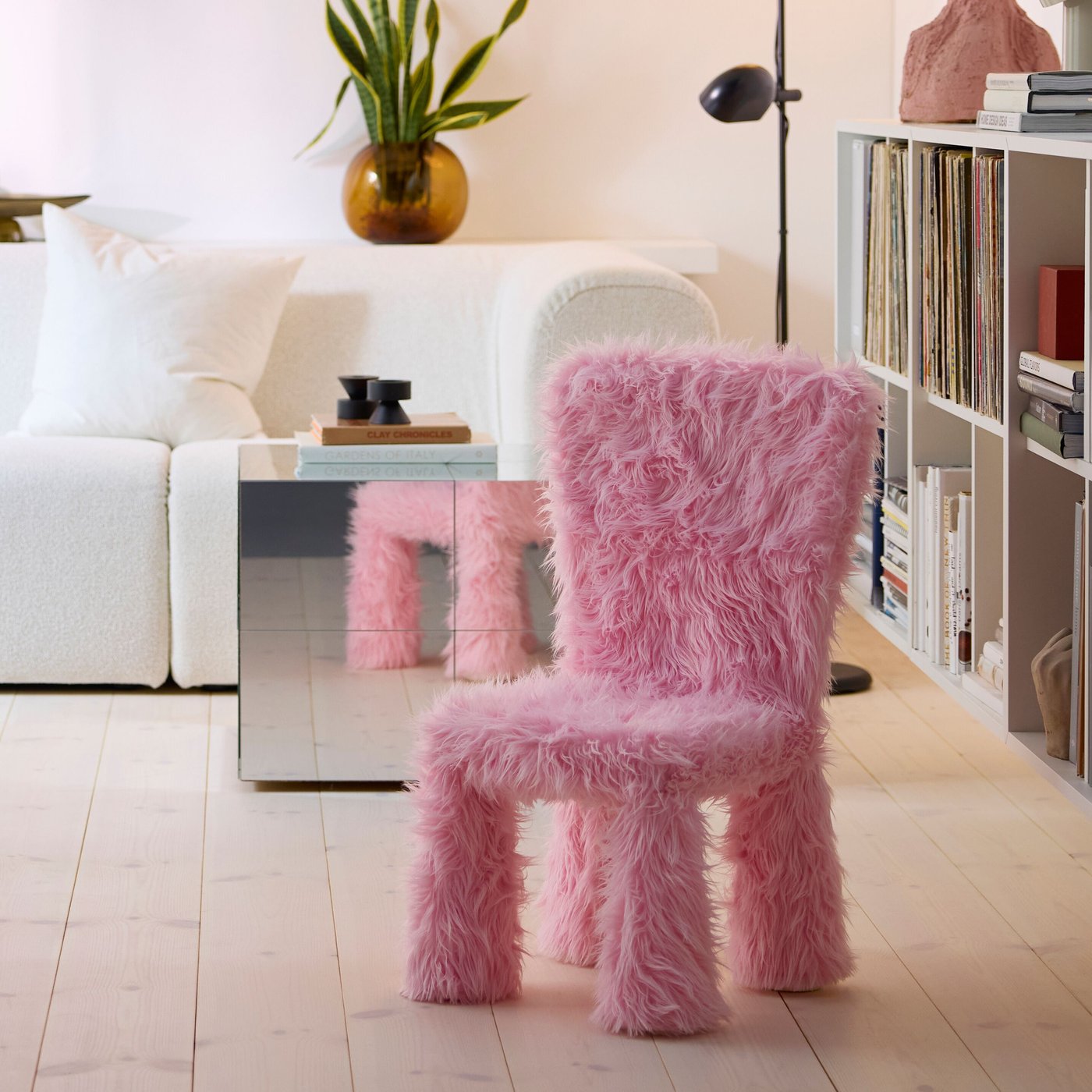 En stol täckt i GREJSIMOJS fluffig rosa textil står i ett modernt vardagsrum. Bakom stolen syns en ljus soffa, ett spegelglänsande sidobord, en hylla fylld med böcker och vinyler samt en växt i en brun vas.