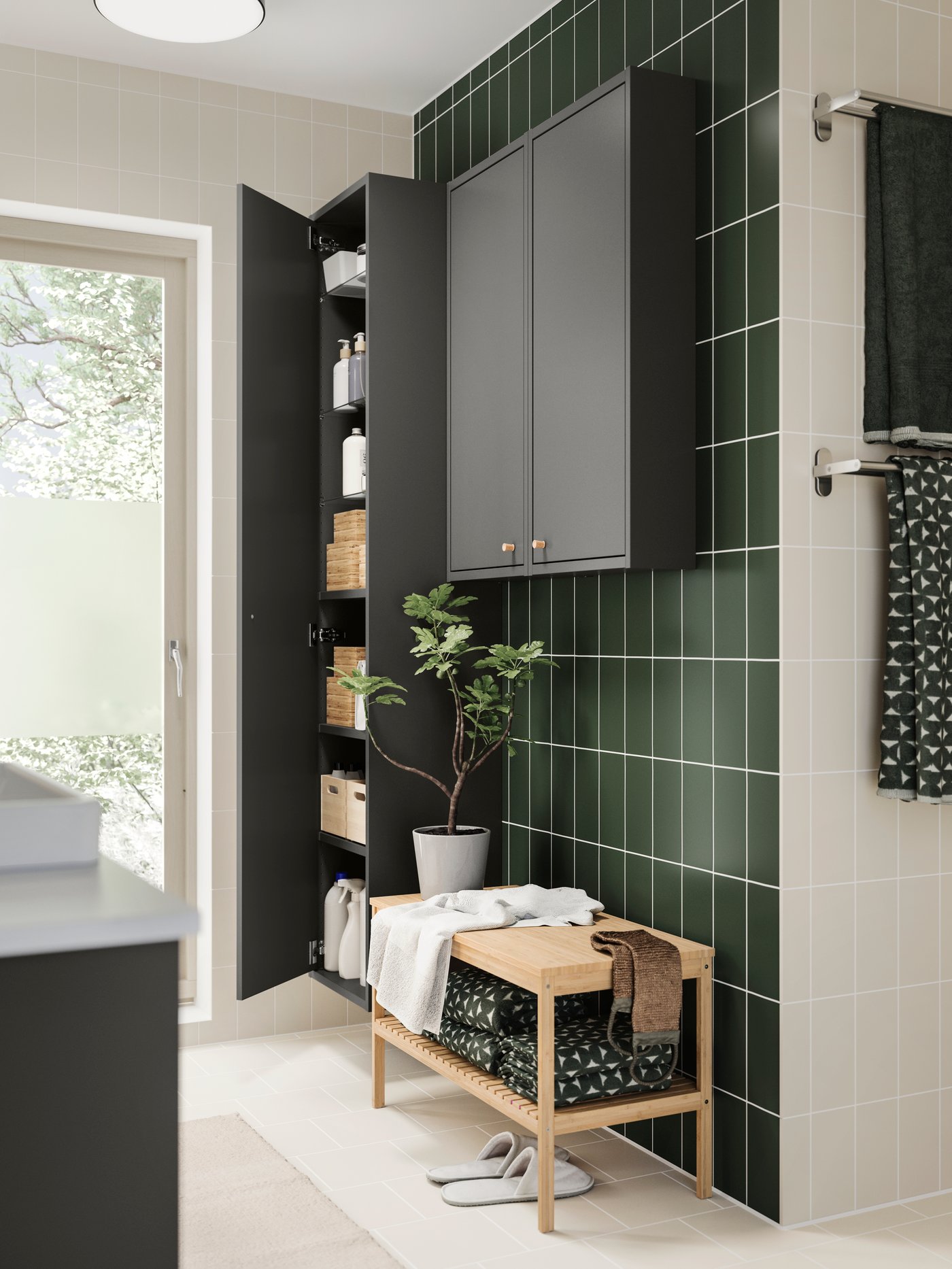 A RÅGRUND bench holding towels and a basket, in front of a tall storage cabinet in a green-tiled bathroom.
