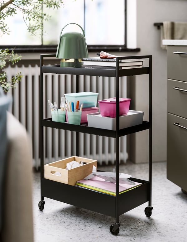 A black NISSAFORS trolley holding a green VAPPEBY Bluetooth speaker lamp, books, pens in mugs, coloured paper and a soft toy.