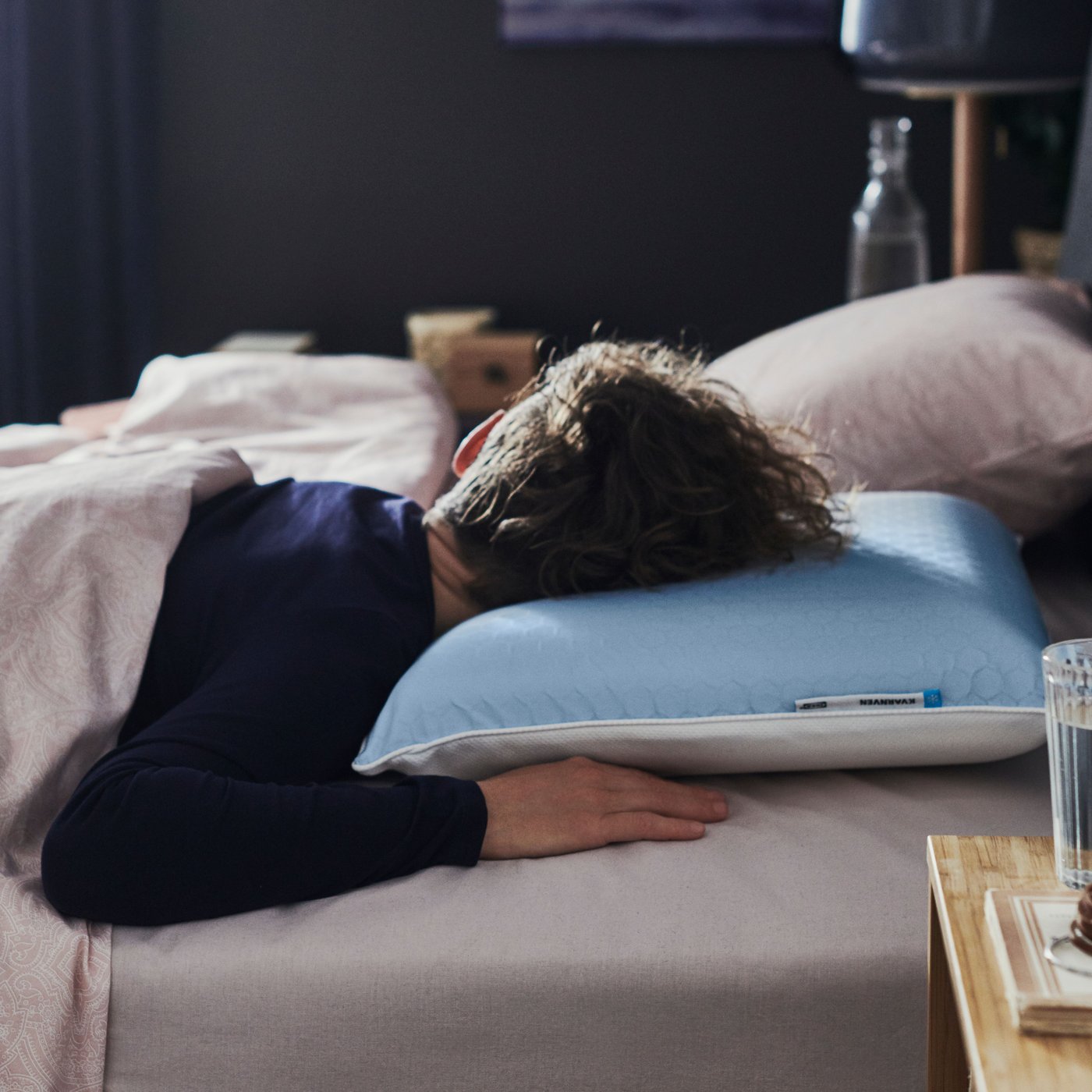 In a dark bedroom, a person in blue pyjamas in bed with a KVARNVEN ergonomic pillow is sleeping on their stomach.