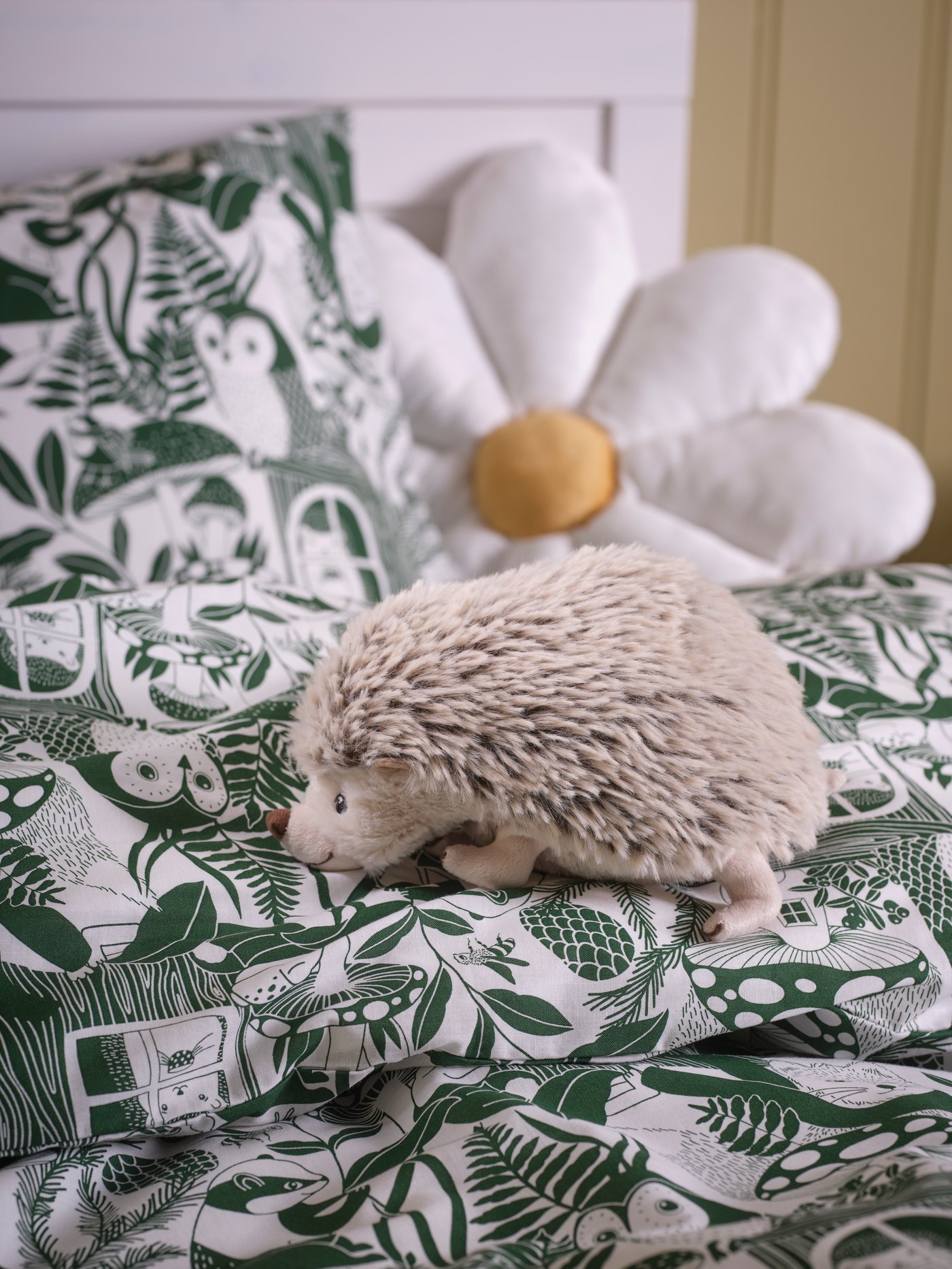 Cojín de margarita SKOGSDUVA sobre una cama y junto a cojín de puercoespín SKOGSDUVA.