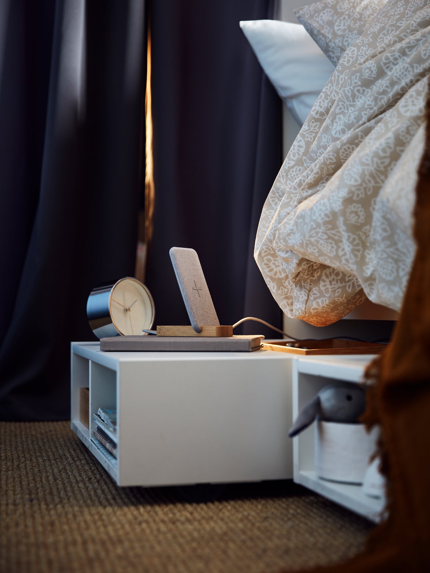 שתי יחידות אחסון/שידות לצד מיטה FREDVANG לבנות הנכנסות מתחת למיטה ומחזיקות ספרים ומגוון פריטים אחרים על שטיח יוטה מתחת למיטה.