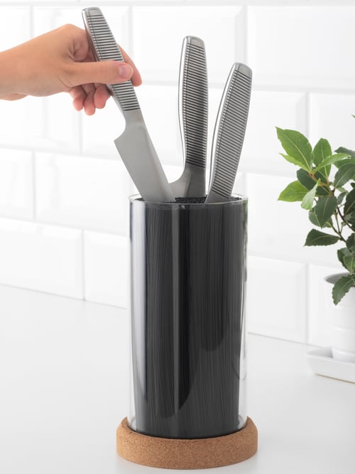 A hand taking out a knife from a black knife block with a cork base, on a white countertop with a white tiled background.