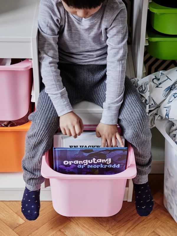 Niño sentado sobre mueble de almacenamiento blanco con rosado TROFAST y libros en su interior.