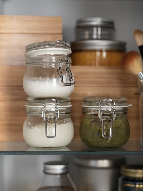 Bucătărie cu borcane de sticlă cu capace și cutii cu capace, conținând ingrediente, așezate pe raftul de sticlă, lângă cutii de lemn.